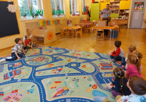 Dzieci siedzą na dywanie w kolorowe wzory. Prowadzący zajęcia objaśnia im, w jaki sposób będą wykonywały świece z wosku pszczelego.