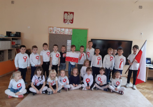Dzieci ubrane na galowo. Dziewczynki siedzą na beżowym dywanie, za nimi stoją chłopcy. Chłopiec z prawej strony trzyma biało-czerwoną flagę Polski.