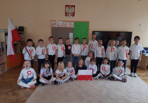 Dziewczynki siedzą na beżowym dywanie, chłopcy stoją za dziewczynkami. Wszyscy są ubrani w stroje galowe. Chłopiec stojący z lewej strony trzyma biało-czerwoną flagę Polski. Dziewczynka siedząca pośrodku trzyma wykonany przez dzieci plakat z flagą Polski.