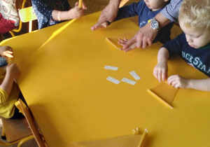 Na zdjęciu chłopiec siedzący z dziećmi przy stoliku, który wykonuje wraz z innymi dziećmi świeczki, chłopcu pomaga organizator warsztatów