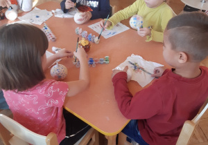 Przy stoliku siedzą trzy dziewczynki i dwóch chłopców. Malują farbami wzory na ceramicznych bombkach.