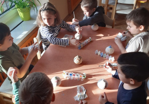 Pięciu chłopców i jedna dziewczynka siedzą przy stoliku i malują farbami ceramiczne bombki.