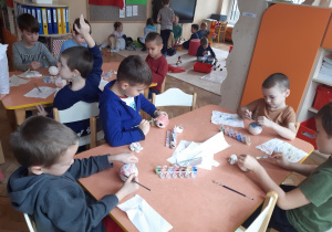 Przy dwóch stolikach siedzi ośmiu chłopców, którzy malują farbami bombki z ceramiki. W tle widoczne są dzieci, bawiące się klockami na beżowym dywanie.