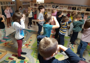 Dzieci stoją na kolorowym dywanie. Naśladują ruchy pani prowadzącej zajęcia- trzymają ręce przy oczach imitując spoglądanie przez okulary. W tle widoczne są szafki na ubrania.