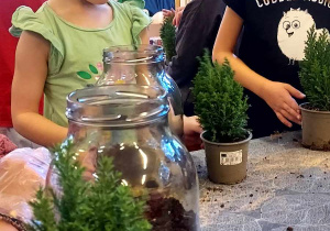 Na zdjeciu dwie dziewczynki stoją przy stole obok słoików i roślin dziewczynki wykonują las w słoiku