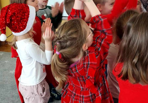 Na zdjęciu dzieci ubrane w w czerwone świąteczne stroje, dzieci stoją na dywanie i wspólnie śpiewają i klaszczą