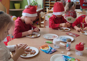 Na zdjęciu dzieci z grupy zielonej siedzą przy stołach i dekorują pierniczki, dzieci ubrane są w stroje koloru czerwonego o tematyce świątecznej