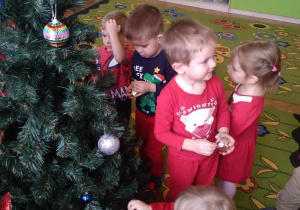 Na zdjęciu dzieci stoją wokół choinki i ją dekorują bombkami, dzieci są ubrane na czerwono