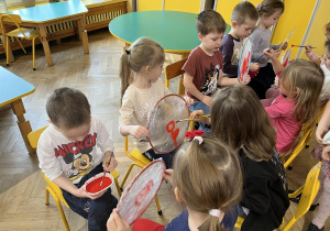 Na zdjęciu dzieci z grupy fioletowej siedzą na krzesełkach na wprost siebie i malują kółeczka oblepione folią, na której namalowane są serduszka