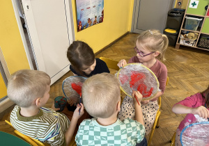 Na zdjęciu dzieci z grupy fioletowej siedzą na krzesełkach na wprost siebie i malują kółeczka oblepione folią, na której dzieci malują serduszka