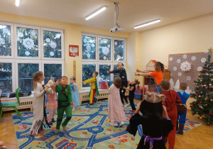 Dzieci i dwie Panie przebrani w karnawałowe stroje tańczą w rytm muzyki. Na szybach okiennych zawieszona jest zimowa dekoracja.