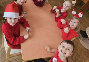 Dzieci ubrane na czerwono siedzą przy stoliku. W rękach trzymają własnoręcznie zrobione i ozdobione pierniczki.