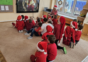Dzieci ubrane na czerwono siedzą na beżowym dywanie. Na ekranie interaktywnym oglądają film z przesłaniem dla nich od Świętego Mikołaja.