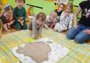 Na zdjęciu dzieci z grupy żółtej siedzą wokół piasku kinetycznego, dzieci odkopują ukryte szczątki dinozaurów