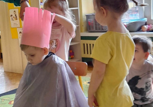 Na zdjęciu dzieci bawią się w fryzjera chłopczyk ma na głowie wysoką czapkę z papieru pozostałe dzieci ćwiczą wycinanie fryzury z papieru
