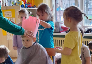 Na zdjęciu dzieci bawią się w fryzjera chłopczyk ma na głowie wysoką czapkę z papieru pozostałe dzieci ćwiczą wycinanie fryzury z papieru