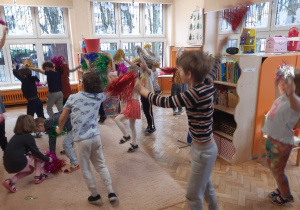 Dzieci ustawione są w rozsypce, w rękach trzymają kolorowe pompony. Dzieci podskakują na beżowym dywanie w rytm muzyki.