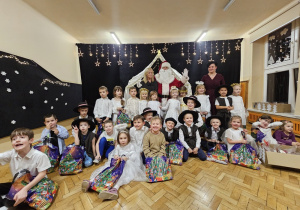Zdjęcie grupowe ze Świętym Mikołajem- dziesięcioro dzieci stoi przed Mikołajem w rzędzie, pozostałe dzieci siedzą na podłodze, w rękach trzymają świąteczne reklamówki z prezentami. Po obu stronach Mikołaja stoją nauczycielki grupy.