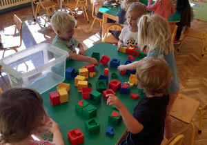 Przy zielonym stoliku siedza trzy dziewczynki i dwóch chłopców. Dzieci bawią się plastikowymi klockami- dopasowują do siebie pasujące elementy.