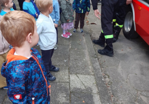 Dzieci stoją w rozsypce. Obok dzieci stoi dwóch strażaków. Jeden chłopiec ma na głowie kask strażacki.