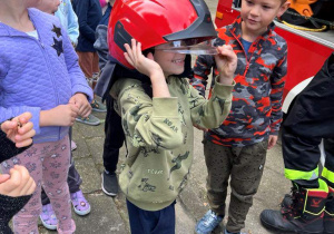 Na zdjęciu chłopiec z hełmem strażacki na głowie chłopczyk stoi obok wozu strażackiego