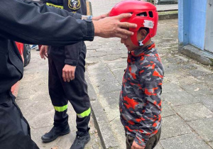 Na zdjęciu chłopiec z hełmem strażacki na głowie, hełm zakłada strażak