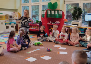 Na zdjęciach dzieci z grupy zielonej dzieci siedzą na dywanie i oglądają prezentowaną przez pedagoga książkę