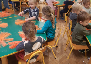 Dzieci siedzą przy stolikach, na których znajdują się wiązki pomarańczowych sznurków.