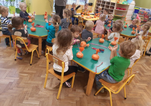 Dzieci siedzą przy stolikach, trzymają w rękach kolorowe sznurki. Na stolikach leżą dynie zrobione z pomarańczowego sznurka oraz kolorowe szczotki do włosów.