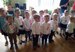 Na zdjęciu dzieci z grupy pomarańczowej , dzieci stoją w grupie zielonej w odświętnych strojach na piersi mają białe czerwone symbole