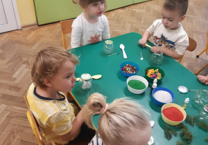 Dwie dziewczynki i dwóch chłopców siedzi przy zielonym stoliku. Na stoliku stoją: słoiczki, miseczki z kolorowym piaskiem, kolorowe ozdoby i gałązki świerkowe.