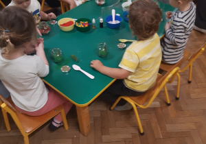 Dzieci siedzą przy stolikach. Wkładają do swoich słoiczków kolorowe ozdoby.