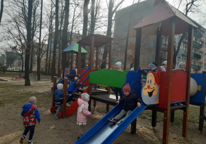 Jeden chłopiec zjeżdża ze zjeżdżalni, dwoje dzieci stoi przed zjeżdżalnią. Troje dzieci wchodzi schodami na zjeżdżalnię. Kilkoro dzieci stoi na podeście zjeżdżalni.