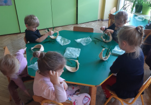 Cztery dziewczynki i jeden chłopiec siedzą przy zielonym stoliku. Wkładają zielone gałązki do beżowych koszyczków ze sznurka.
