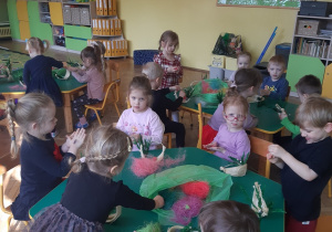 Dzieci siedzą przy zielonych stolikach. Z kolorowego sizalu robią kuleczki. W tle widać żółtą ścianę oraz beżową tablicę, na której zawieszona jest wiosenna dekoracja.