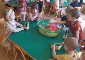 Dzieci siedzą przy zielonych stolikach. Na stolikach leży kolorowy sizal. W tle widać zieloną szafę, żółtą ścianę i białe drzwi. Na szafie wisi plakat z kolorowymi odciskami dłoni dzieci.