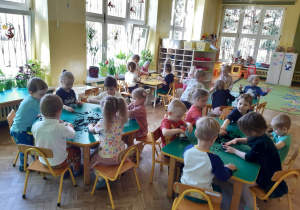 Dzieci siedzą przy trzech zielonych i jednym żółtym stole. Na stołach znajduje się rozsypana ziemia i przezroczyste pojemniki. W tle widać okna i brązową szafkę z półkami.