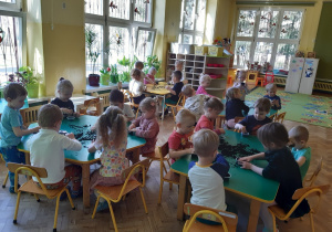 Dzieci siedzą przy trzech zielonych i jednym żółtym stole. Na stołach znajduje się rozsypana ziemia i przezroczyste pojemniki. W tle widać okna i brązową szafkę z półkami.