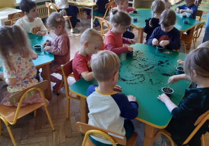 Dzieci siedzą przy trzech zielonych i jednym żółtym stole. Na stołach znajduje się resztka czarnej ziemi, pojemniki z ziemią i nasiona owsa. W tle widać okna i zielone kwiaty w zielonych doniczkach postawione na białym parapecie oraz brązową szafkę z półkami.