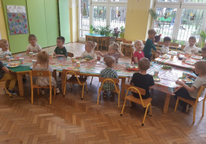 Dzieci siedzą przy złączonych stołach okrytych kolorowym, świątecznym obrusem. Na stole stoją kubki i talerzyki ze świątecznymi motywami. Na półmiskach ułożone jest jedzenie. W tle widać okna, na których przywieszone są wiosenne dekoracje.