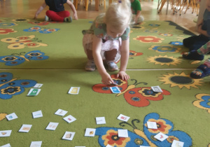 Dzieci siedzą na brzegu zielonego dywanu w kolorowe motyle i kwiaty. Jedna dziewczynka kuca na środku dywanu układając obrazki ze śmieciami przy obrazkach odpowiednich koszy na śmieci. W tle widać brązowe krzesełka, żółtą ścianę oraz zieloną szafę.