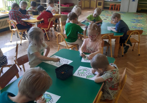 Dzieci siedzą przy dwóch zielonych i jednym żółtym stoliku. Na stolikach w czarnych pudełkach stoją kredki. Dzieci kolorują kwiatka z środkiem przypominającym kulę ziemską. W tle widać okna, brązową szafkę z półkami oraz żółte mebelki z kącika lalek.
