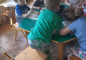Dzieci siedzą przy zielonym i żółtym stoliku. Na stolikach w czarnych pudełkach stoją kredki. Dzieci kolorują kwiatka z środkiem przypominającym kulę ziemską.