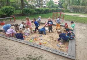 Dzieci bawią się w piaskownicy kolorowymi, plastikowymi zabawkami. W tle widać czerwone drabinki. Po lewej stronie stoi brązowo-czerwona ławka.
