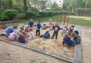 Dzieci bawią się w piaskownicy kolorowymi, plastikowymi zabawkami. W tle widać czerwone drabinki. Po lewej stronie stoi brązowo-czerwona ławka oraz zielone krzaki i brązowe ogrodzenie.