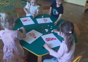 Trzy dziewczynki i jeden chłopiec siedzą przy zielonym stoliku. W rękach trzymają kleje. Na stoliku leżą kartki z konturami mapy Polski podzielonej na pół w kolorze biało-czerwonym oraz białe i czerwone papierowe koła. W tle widać żółte segregatory ustawione na brązowej szafce, żółtą ścianę oraz brązowe szafki z żółtymi i zielonymi frontami.
