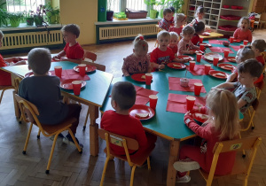 Dzieci ubrane na czerwono siedzą przy połączonych stolikach. Na stołach znajdują się czerwone talerzyki, kubki i serwetki oraz półmiski z jedzeniem w odcieniach czerwieni.