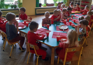 Dzieci ubrane na czerwono siedzą przy połączonych stolikach. Na stołach znajdują się czerwone talerzyki, kubki i serwetki oraz półmiski z jedzeniem w odcieniach czerwieni.