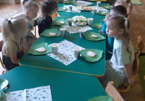 Dzieci ubrane na zielono siedzą przy połączonych stolikach. Na stołach znajdują się zielone talerzyki w kształcie liścia, zielone kubki i serwetki oraz półmiski z jedzeniem w odcieniach zieleni.