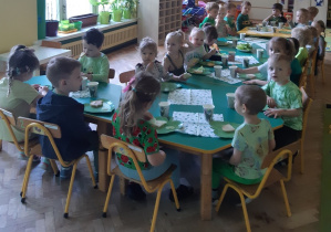 Dzieci ubrane na zielono siedzą przy połączonych stolikach. Na stołach znajdują się zielone talerzyki w kształcie liścia, zielone kubki i serwetki oraz półmiski z jedzeniem w odcieniach zieleni.